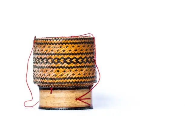 A sticky rice container, made of bamboo strips woven into a pattern and shaped of a cylinder. This basketry has two parts, the container and the cover tying together by the red rope.