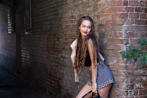 Portrait of a teenage girl in a youth style standing by a brick wall. In a black top and short skirt.