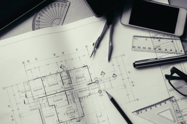 istock Divider, pencil, pen, ruler, glasses and smartphone and blueprint on table top.Table top view of Engineers table at office workplace.selective focus.Man hand point to blueprint.Black and white effect. 824913896