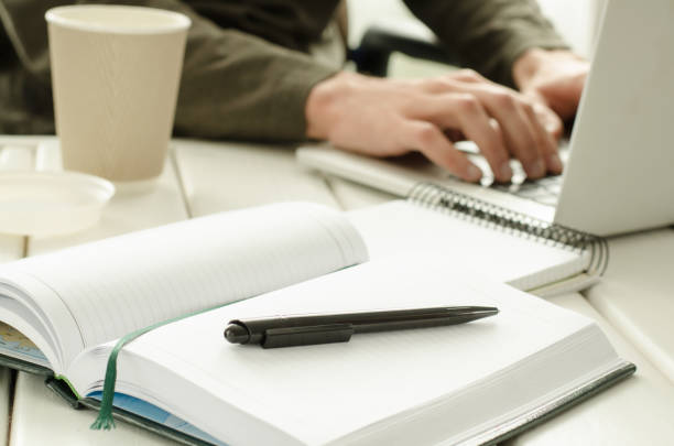 istock Open notepad with black pen on work-table. Paper cup of coffee, office stuff, laptop and working man on background 824924050
