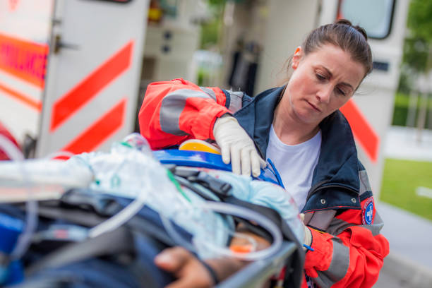 Female paramedic with patient Front close up view of a female doctor helping unrecognizable injured person with head protector lying on stretcher, ambulance vehicle in background. medic stock pictures, royalty-free photos & images