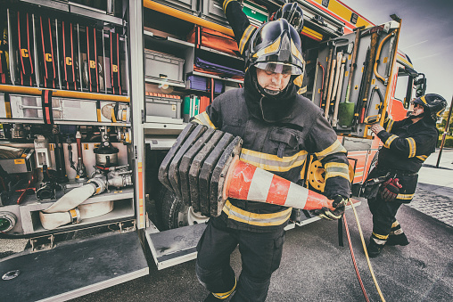 Close up of two fire fighters in front of a fire engine, one is holding traffic cones.