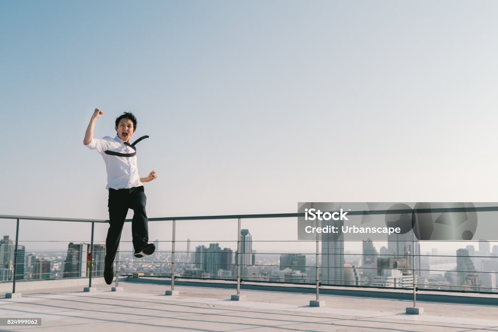 Young handsome Asian businessman jumping high, celebrate success winning pose on building rooftop. Work, job, or successful business concept. Cityscape background with copy space on sunny blue sky Jumping Stock Photo
