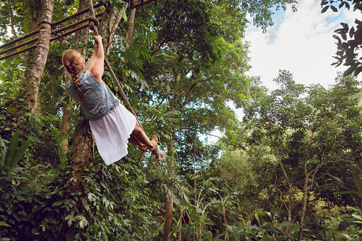 Family travel lifestyle. Happy young woman flying high with fun on rope swing on wild jungle background. Funny adventure walk in tropical rainforest. Leisure activity on summer vacation with kids.
