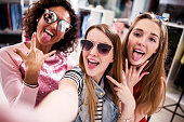 Pretty girls wearing sunglasses fooling around taking selfie showing tongue and horn gestures in clothing shop