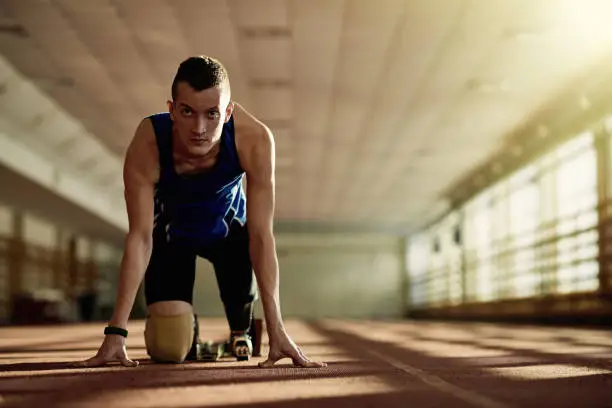 Motivational portrait of handicapped young athlete with prosthetic leg in start position on running track during sport practice