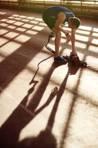 Portrait of adaptive athlete with prosthetic leg setting starting blocks on running track during sport practice lit by sunlight