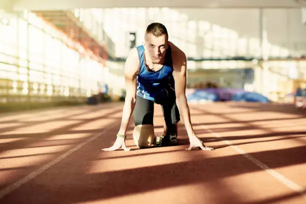 Portrait of strong willed athlete with prosthetic leg in start position on running track, looking confidently at camera
