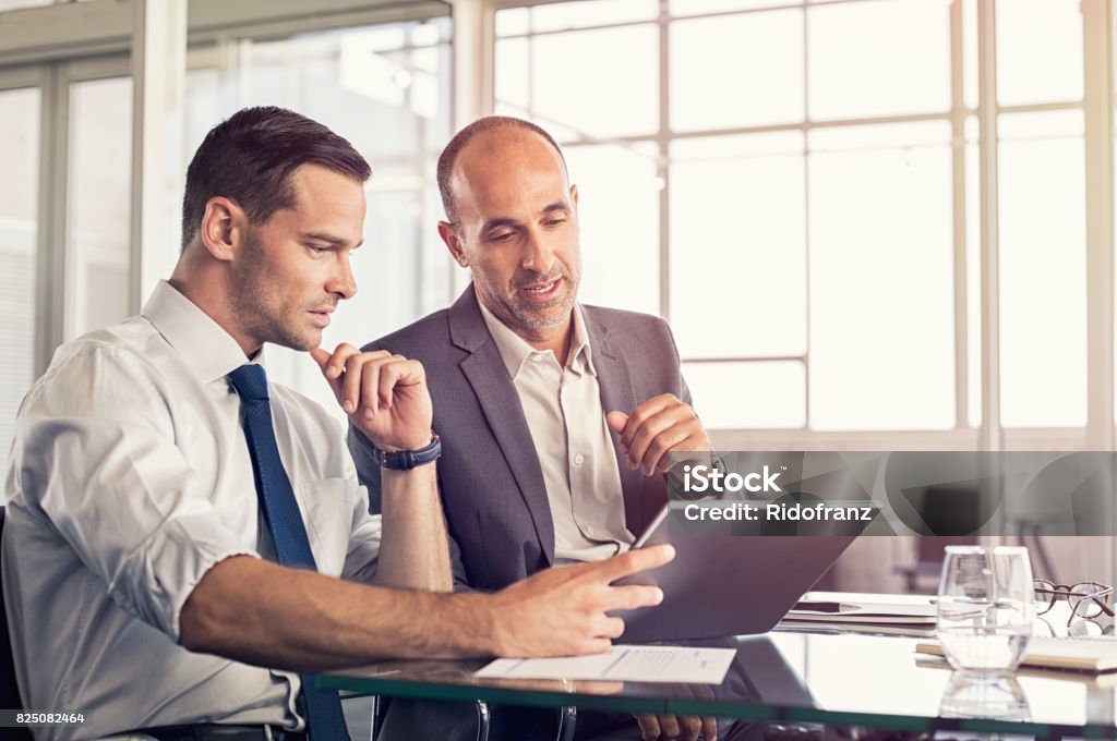 Businessmen working on digital tablet Businessman using a digital tablet to discuss information with senior leadership in a meeting. Business partners discussing plans. Mature boss and young business man working together in office. Businessman Stock Photo