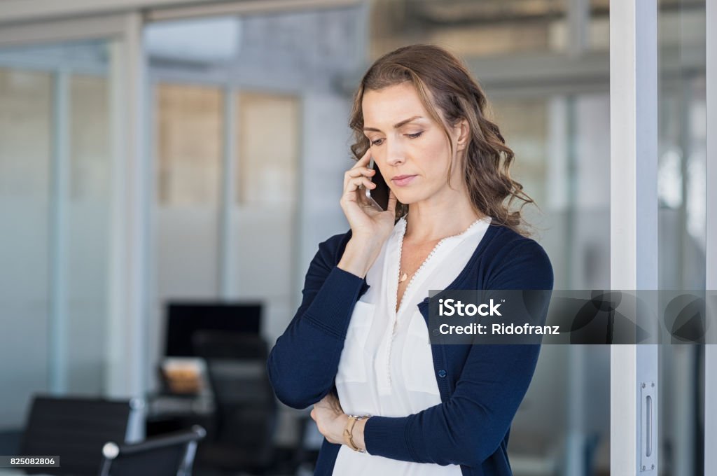 Serious businesswoman talking on phone Serious business woman looking worried while talking on the phone in conference room. Frustrated businesswoman in office using mobile phone. Unhappy mature woman talking on phone call on smartphone. Using Phone Stock Photo