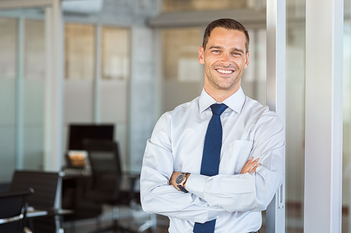 Portrait of cheerful businessman with arms folded standing in conference room. Happy young business man in shirt and tie looking at camera. Portrait of a smiling businessman in modern office with copy space.