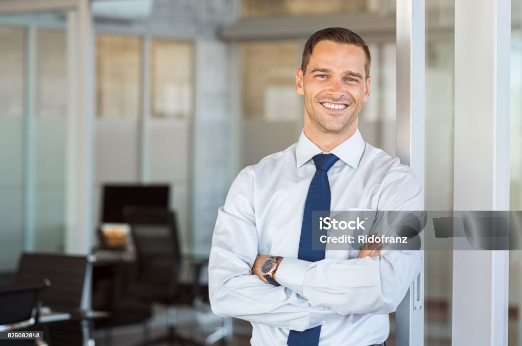 Smiling businessman at office Portrait of cheerful businessman with arms folded standing in conference room. Happy young business man in shirt and tie looking at camera. Portrait of a smiling businessman in modern office with copy space. Men Stock Photo