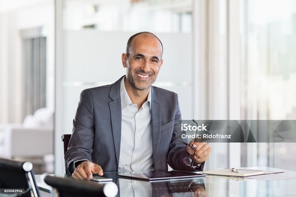 Happy mature business man Happy mature business man holding spectacles in modern office. Successful senior businessman in formals sitting in meeting room with phone and tablet. Smiling man in suit in a elegant office. Financial Advisor Stock Photo