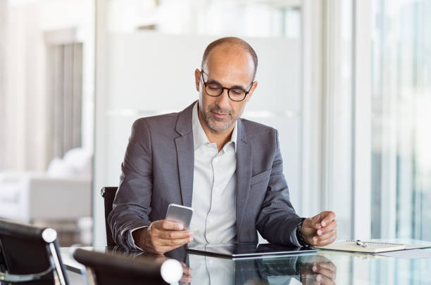 Businessman working on phone Mature business man in formal clothing wearing spectacles using mobile phone. Serious businessman using smartphone and digital tablet at work. Manager in suit using cellphone in a modern office. mature businessman stock pictures, royalty-free photos & images