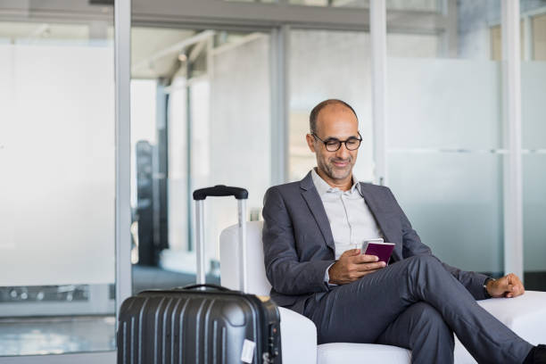 Mature businessman at airport Mature businessman using mobile phone at the airport in the waiting room. Business man typing on smartphone in lounge area. Portrait of latin man sitting and holding passport with luggage. airport departure area stock pictures, royalty-free photos & images