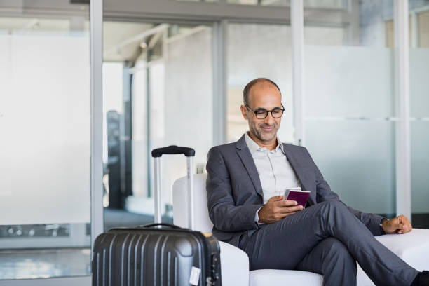 Mature businessman at airport Mature businessman using mobile phone at the airport in the waiting room. Business man typing on smartphone in lounge area. Portrait of latin man sitting and holding passport with luggage. business journey stock pictures, royalty-free photos & images