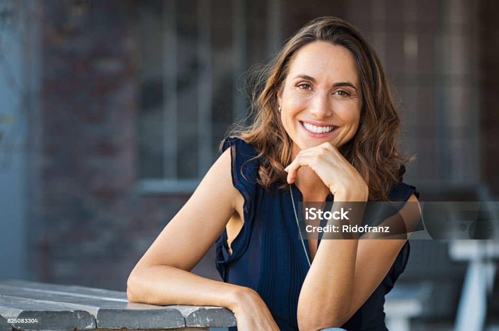 Happy mature woman outdoor Portrait of beautiful mature woman sitting at coffee shop. Happy hispanic smiling woman sitting on a bench in outdoor cafeteria looking at camera. Portrait of carefree woman relaxing on bench. Women Stock Photo