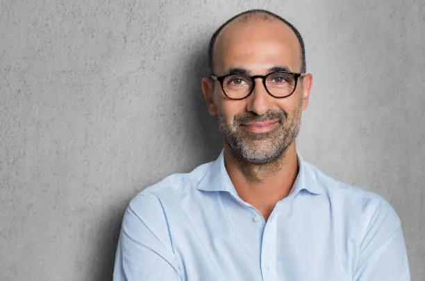 Portrait of a mature businessman wearing glasses on grey background. Happy senior latin man looking at camera isolated over grey wall with copy space. Close up face of happy successful business man.