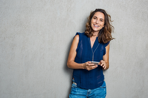 Smiling mature woman using smartphone and looking at camera. Happy woman typing on cellphone over gray background with copy space. Portrait of smiling latin woman messaging with smartphone.