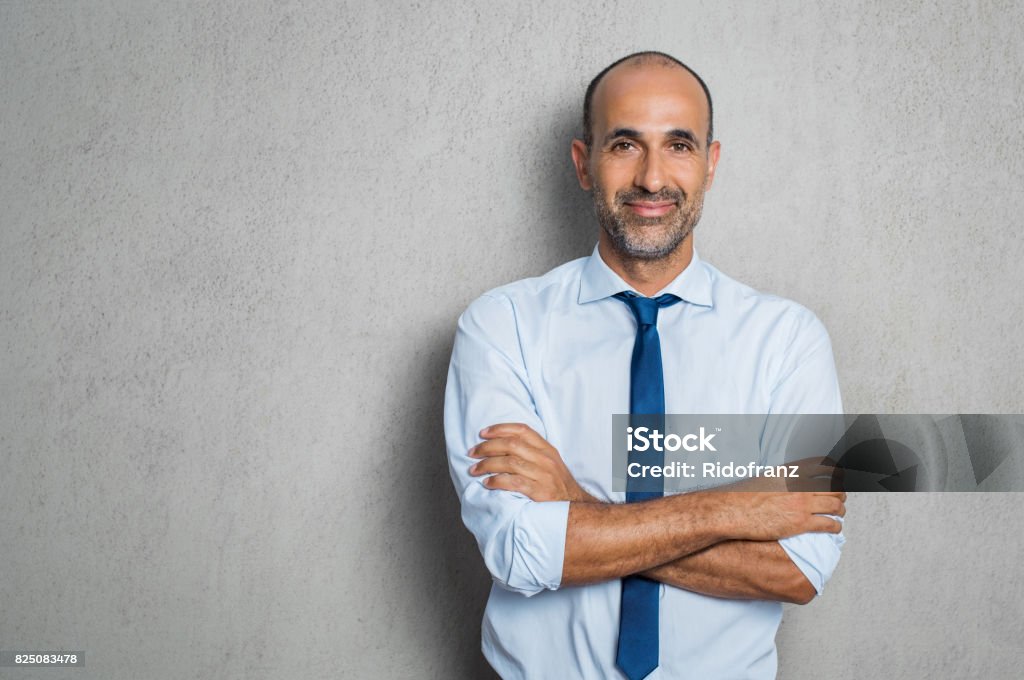 Happy mature businessman Happy mature businessman in blue shirt and tie looking at camera. Portrait of smiling and satisfied hispanic business man with arms crossed isolated over grey background with copy space. Successful senior man standing on grey wall. Men Stock Photo