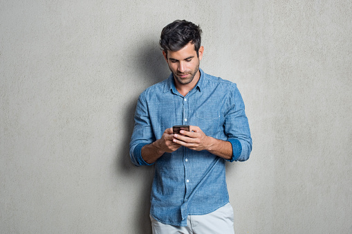 Young man writing phone message. Mid adult man typing on mobile while standing on grey background. Latin man leaning on gray wall while browsing internet on smartphone.