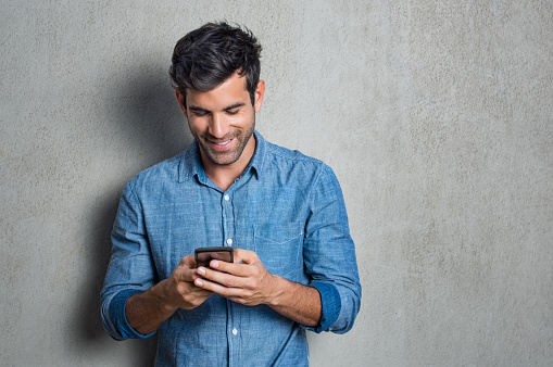 Young man texting message on smart phone isolated on grey background. Smiling latin man holding smartphone and looking at it. Happy hispanic man writing a message on the  phone.