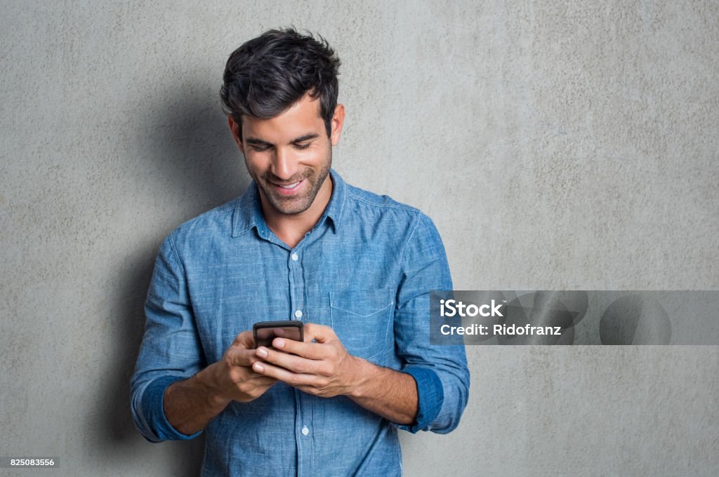 Man texting on phone Young man texting message on smart phone isolated on grey background. Smiling latin man holding smartphone and looking at it. Happy hispanic man writing a message on the  phone. Men Stock Photo