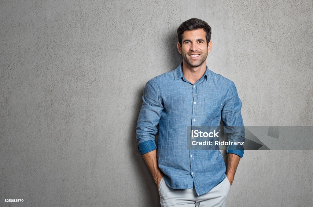 Latin man standing Portrait of a handsome young man smiling against grey background with copy space. Smiling latin guy with hands in pocket in blue shirt standing and leaning on wall. Successful hispanic man looking at camera. Men Stock Photo