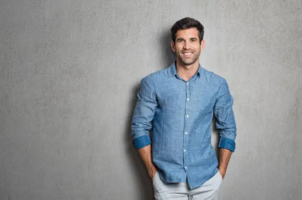 Portrait of a handsome young man smiling against grey background with copy space. Smiling latin guy with hands in pocket in blue shirt standing and leaning on wall. Successful hispanic man looking at camera.