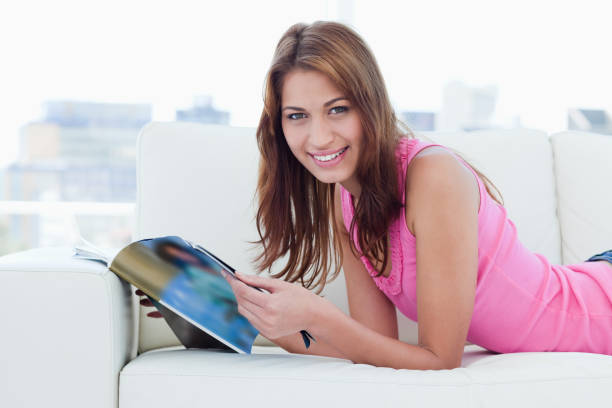 istock Smiling young woman reading a magazine on the sofa while looking at the camera 825100842