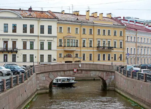 Neo-classical building facades crowd the Winter Canal as apleasure craft navigates a small road bridge spanning it, St.Petersberg streetscene, Russia