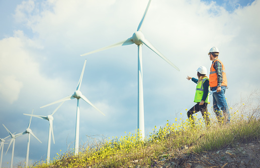 Two young male and female wind farm engineers wearing hi vis jackets and white hardhats standing, working, checking farm field system and looking up at wind turbine farm power generation station in rural landscape. XXXL size