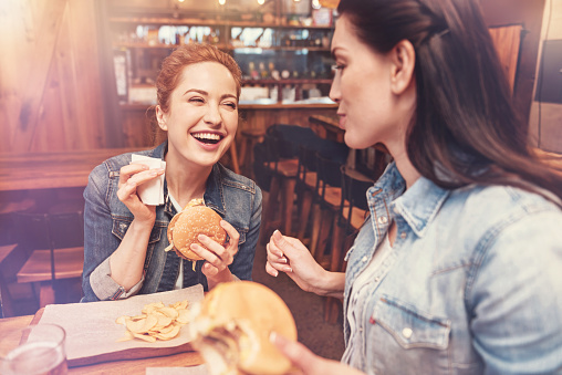 Be positive. Delighted young woman keeping smile on her face and holding burger in left hand while looking aside
