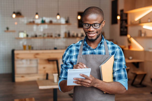 istock handsome smiling african american waiter taking order with notepad and digital tablet in coffee shop 825195324