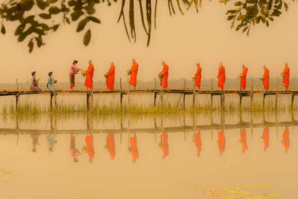 Mahasarakham: Three women in traditional clothing prepare and offer alms to the monks on wooden bridge in the morning with foggy environment in Mahasarakham, Thailand