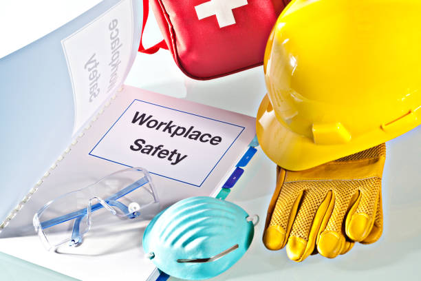 A still life of a Workplace Safety Manual instruction book, with a variety of safety equipment such as hard hat, work gloves, eye gargles breathing dust mask and first aid pack.