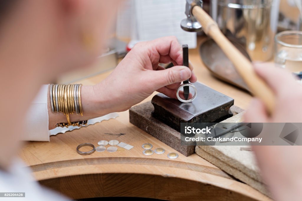 Goldsmith at Work. Portrait of a professional Goldsmith at work in her workshop creating a silver ring. In this picture she is stamping her hallmark on the inside on the ring. She is surrounded by the tools she uses to create her pieces of jewellery. Colour, horizontal with some copy space. Making Stock Photo
