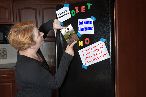 Mature overweight woman posting diet motivational signs and picture of younger, thinner self on kitchen refrigerator.