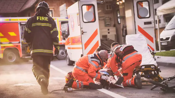 Paramedic team knealing around injured cyclist lying on street, damaged bicycle beside them, firefighter walking past them.