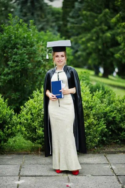 Photo of Portrait of a beautiful female graduate in dress and graduation gown with hat posing in the park.
