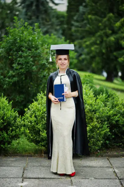 Photo of Portrait of a beautiful female graduate in dress and graduation gown with hat posing in the park.
