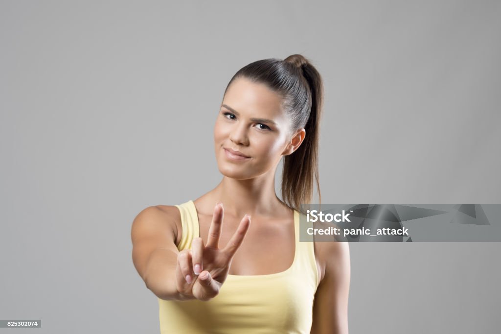 Gorgeous sporty young female portrait showing two finger peace hand sign Gorgeous sporty young female portrait showing two finger peace hand sign over gray studio background. Showing Stock Photo