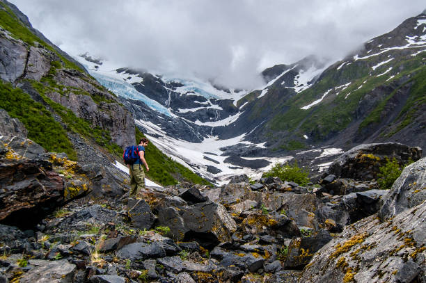 View of Byron glacier and a tourist View of Byron Glacier on the background and massive rocks covered with moss and plants in the foreground with a young man walking up towards the glacier. Shot in the USA, Alaska, in summer portage valley stock pictures, royalty-free photos & images
