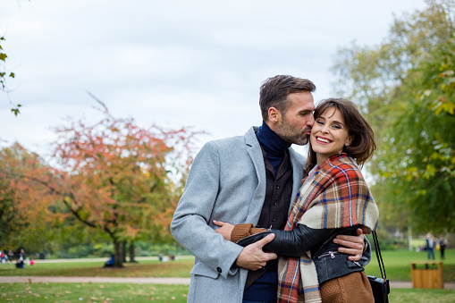 Handsome man kissing his wife on cheeks at the park. Happy couple enjoying themselves at public park on autumn day.