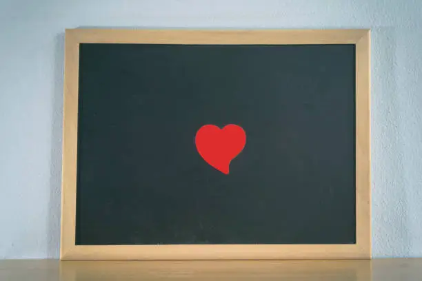 Black board and red-heart shape paper for note message on table with concrete wall background. Love and valentine concept.