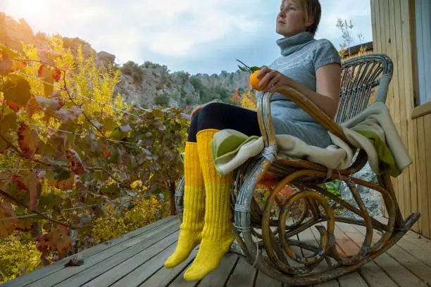 Cute young tall lady in bright yellow knitted socks with a cup in hand admiring the view of the mountains and nature, sitting in a rocking chair. Start of holiday.Ray of sunshine.