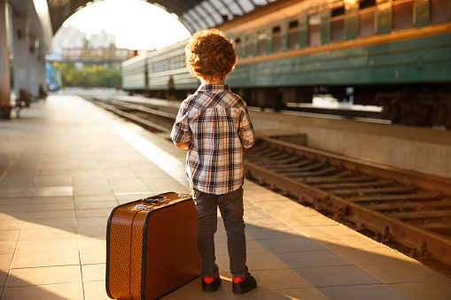 Back view of trendy boy posing on railway station with suitcase.