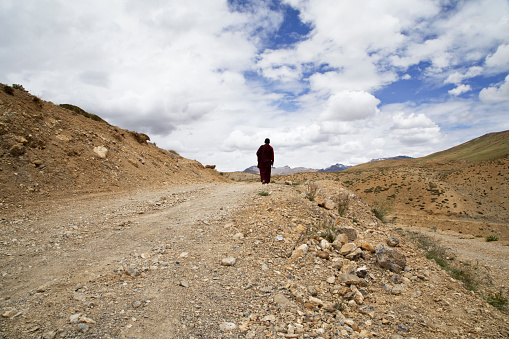 Rear view of monk walking on dirt road against cloudy sky