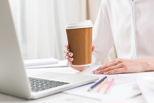 cropped shot of businesswoman holding paper cup while using laptop