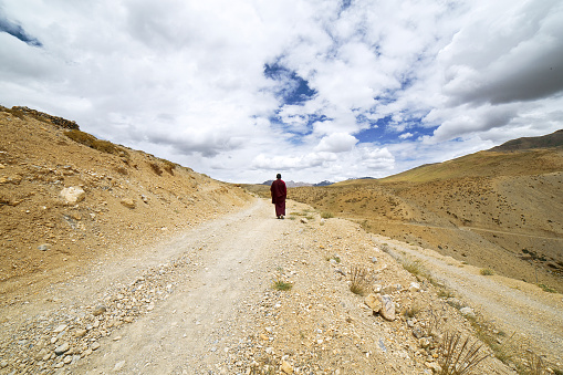 Rear view of monk walking on dirt road against cloudy sky
